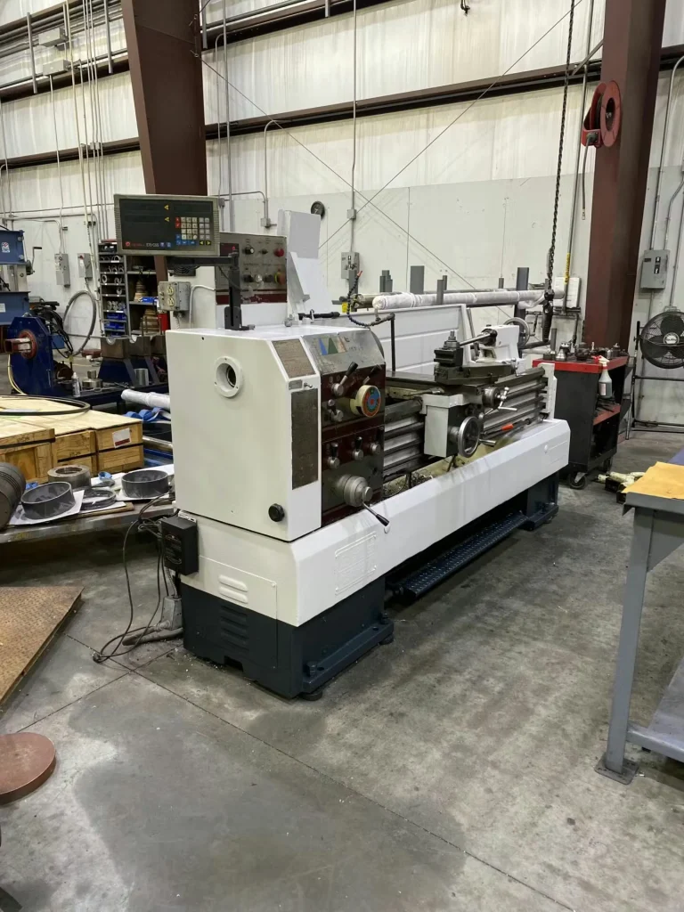 Large lathe in a machine shop, viewed from the headstock end; a white bucket sits on the floor nearby, with shelves, carts, and shop tools around the machine.