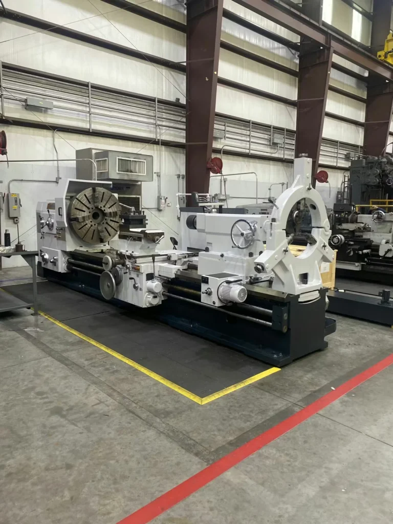 Very large lathe with a prominent steady rest/support frame on the right side and a large faceplate on the left; yellow tape marks a rectangular safety zone on the floor.