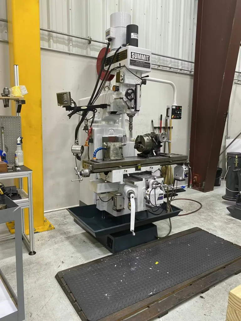 Machine shop area with a white surface grinder and nearby equipment; red floor lines mark a boundary, and electrical panels and conduit run along the wall behind the machines.