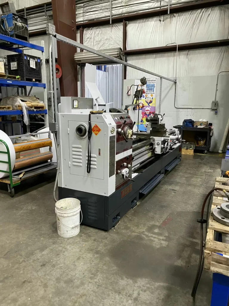 White engine lathe in a workshop with a digital readout above the headstock; tooling, cables, and a worktable surround the machine in a busy shop environment.