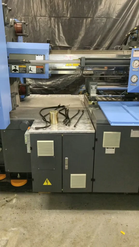 Close view of a large blue industrial machine with metal rollers and warning labels; black hoses and a brass-colored cylinder rest on a diamond-plate surface above gray access panels, with a dark tarp backdrop behind the equipment.