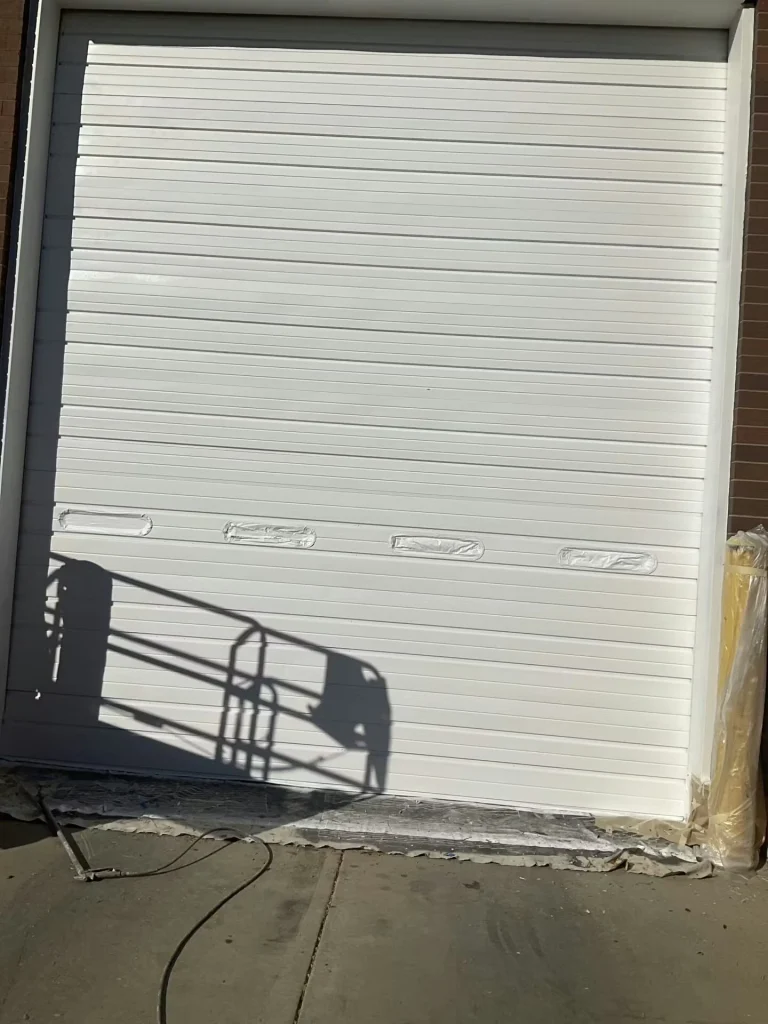 Large white roll-up overhead door with horizontal slats; several small rectangular window openings are masked with plastic, and the shadow of a lift or railing falls across the lower portion.