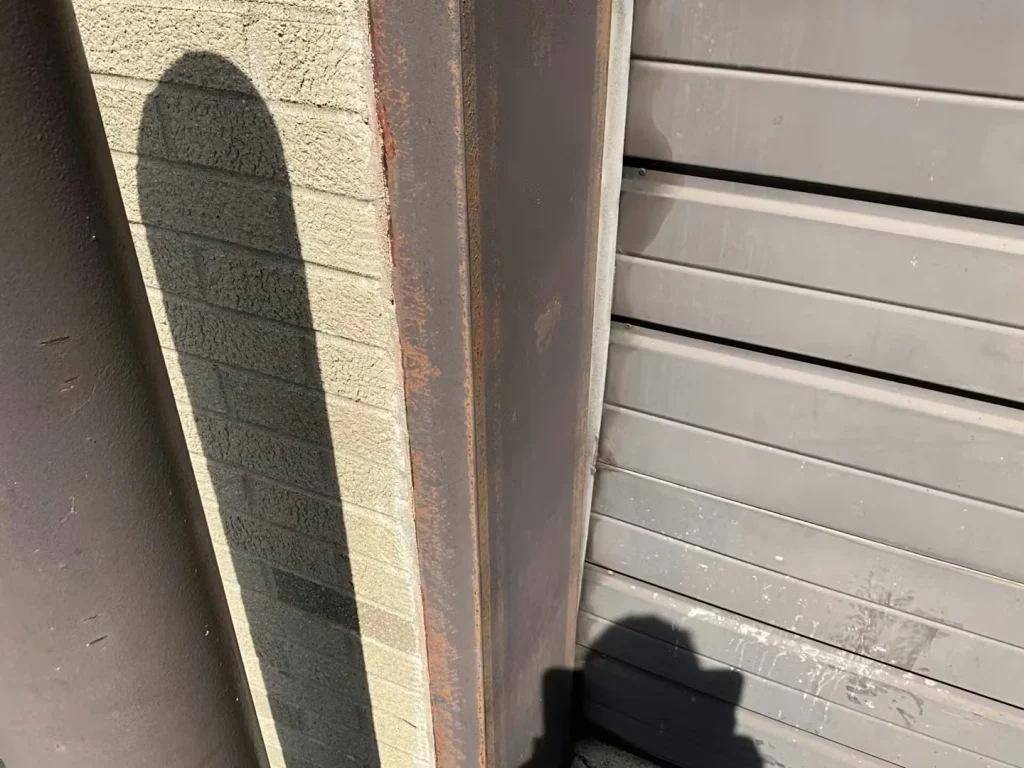 Close-up of the edge of a gray overhead door next to a brown metal frame and textured masonry wall; rust spots are visible on the frame, with strong shadows cast in sunlight.