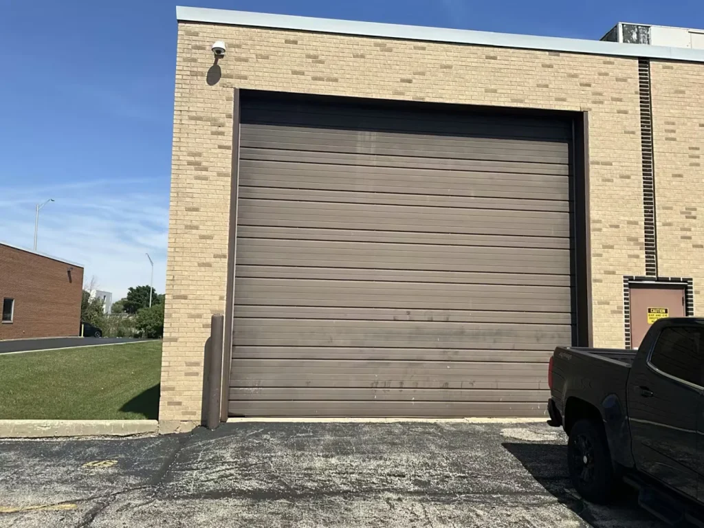 Wide view of a brick industrial building facade with a closed brown overhead door; a pickup truck is parked at the right edge of the frame, with asphalt and grass visible nearby.