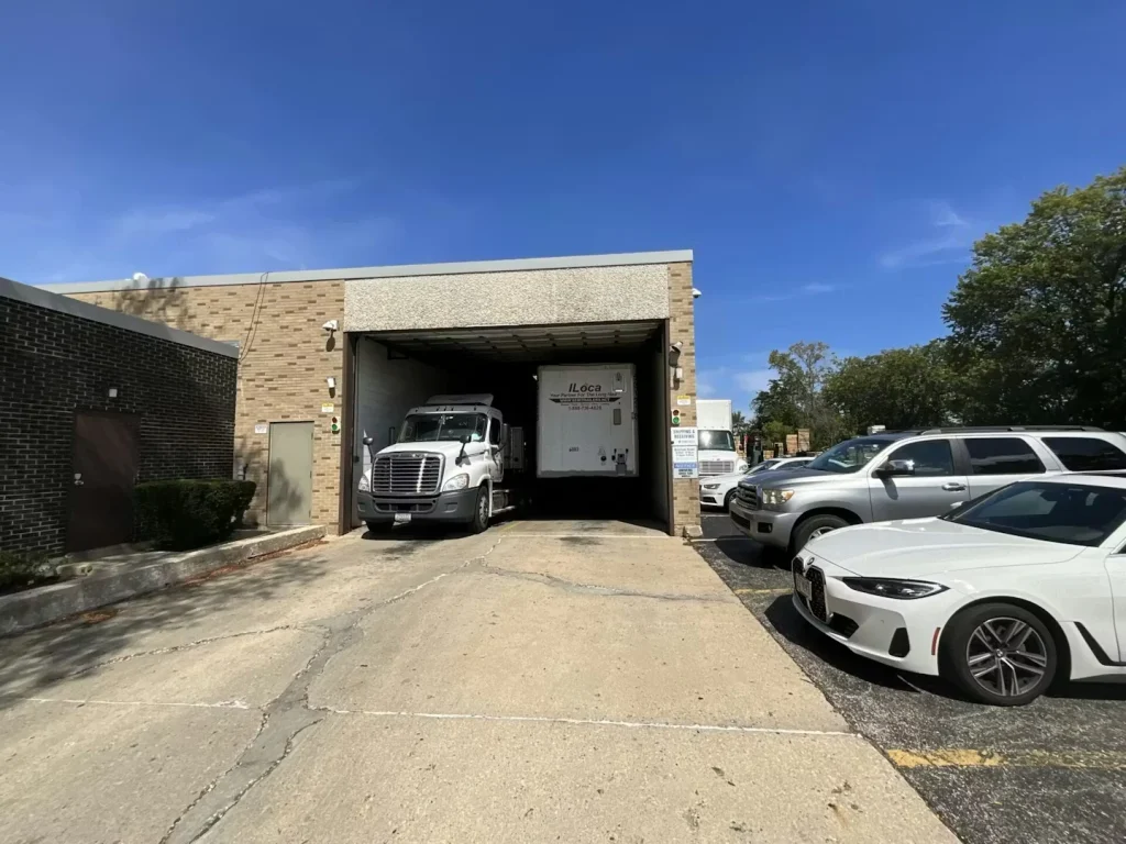 Loading dock entrance with a semi-truck backed into a recessed bay; parked cars line the right side of the driveway under a clear blue sky.