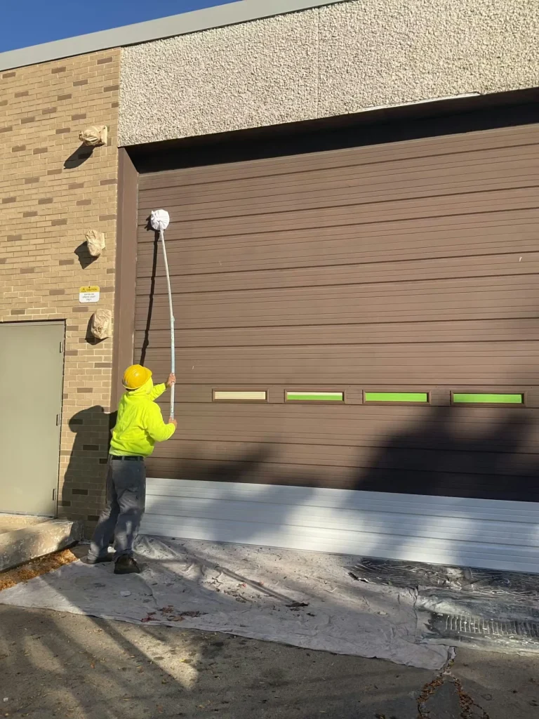 Worker in a yellow hard hat and high-visibility sweatshirt uses a long-handled roller to paint a large brown overhead door; drop cloths cover the ground and a freshly painted white lower section is visible.