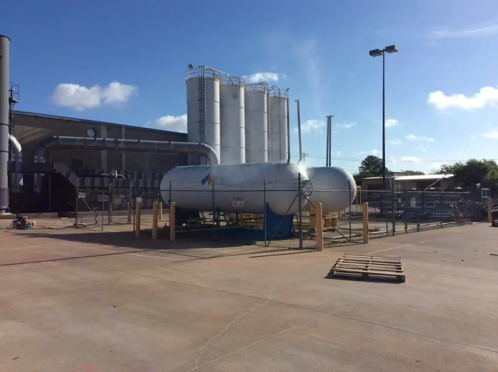 Industrial yard with a large horizontal white storage tank inside a chain-link fenced enclosure; tall white silos and factory piping rise behind it under a bright blue sky, with an empty concrete lot and a wooden pallet in the foreground.