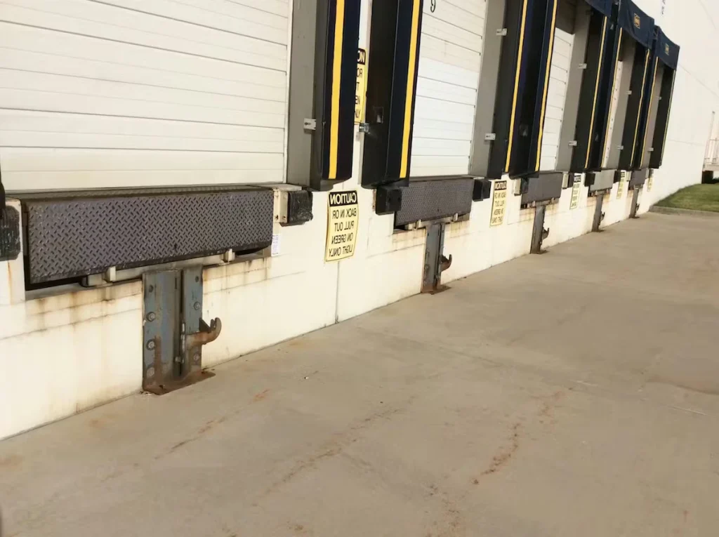 Row of warehouse loading dock doors with black-and-yellow dock bumpers and metal dock levelers; rust staining and grime are visible along the lower wall near the supports, with a wide concrete drive in front.