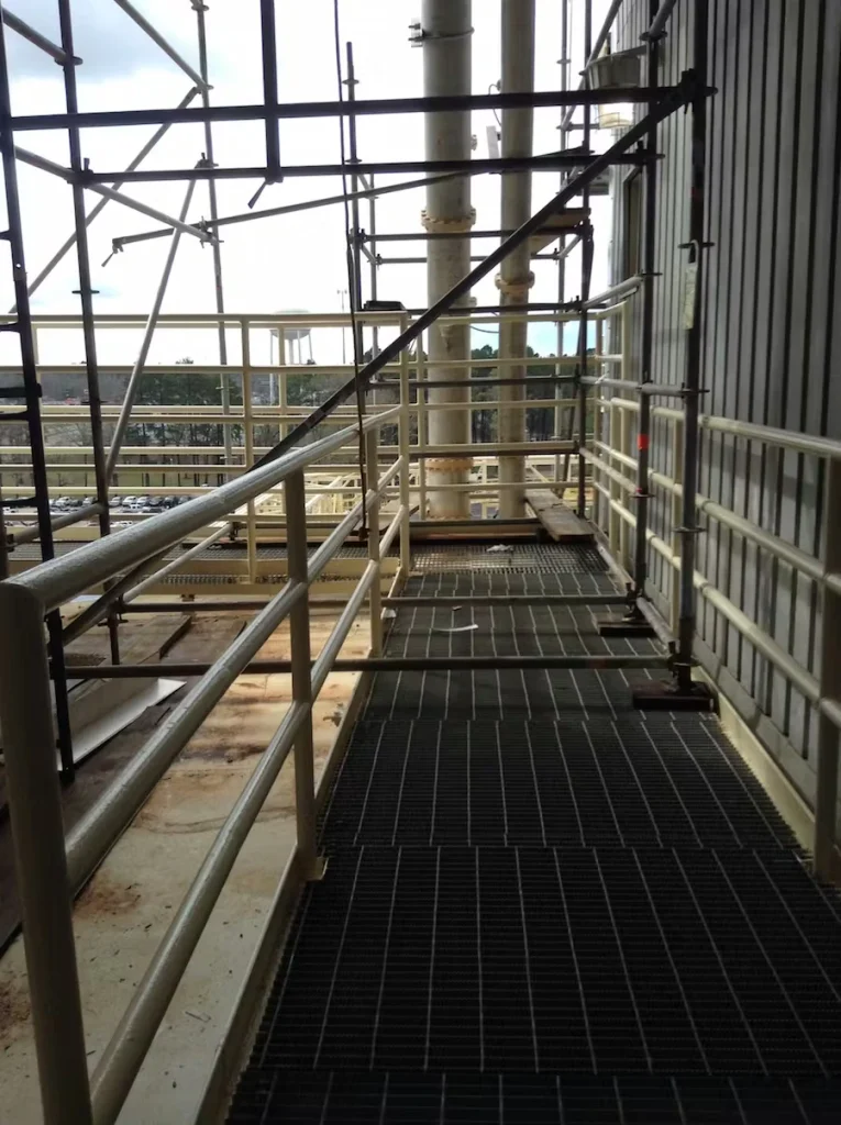 Narrow elevated industrial catwalk with grated flooring and beige guardrails, running beside a corrugated metal wall; scaffolding frames the walkway and tall vertical pipes rise ahead, with the facility yard visible through the railings.