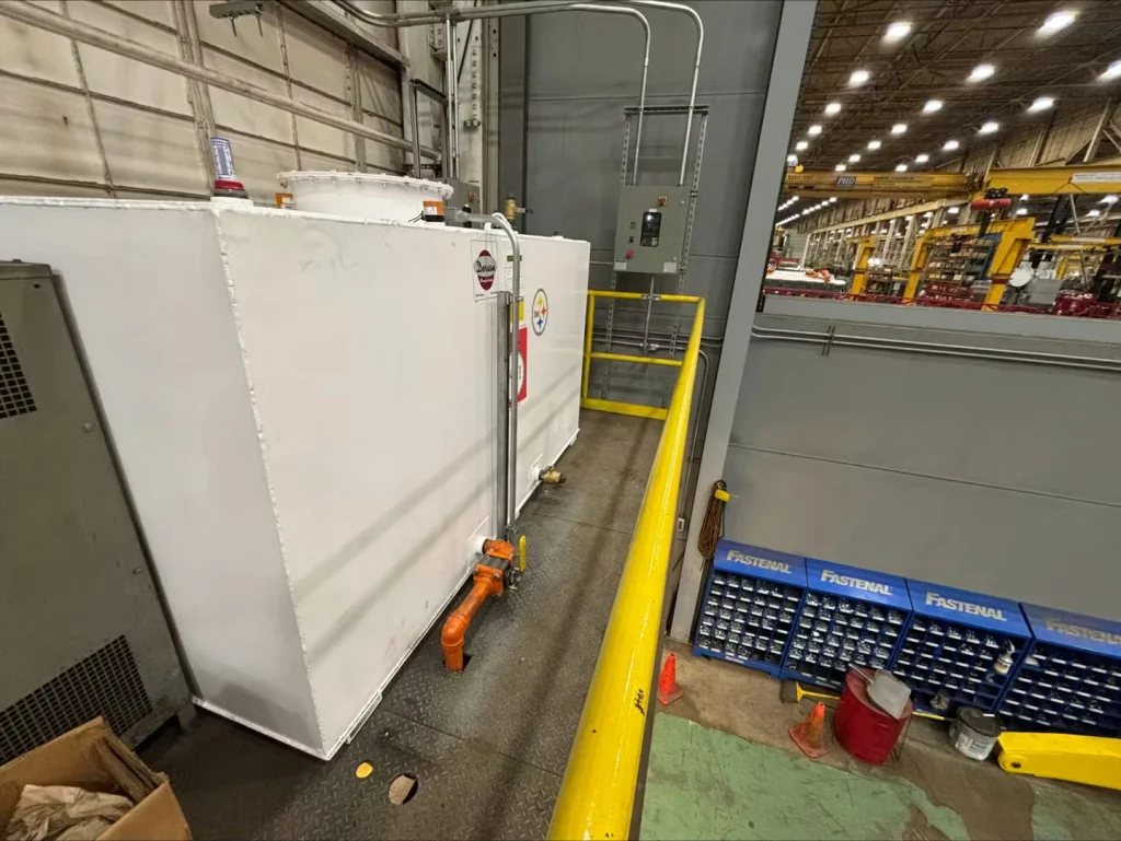 Orange rectangular industrial tank on a factory mezzanine beside a yellow guardrail, with orange piping and valves; machinery and the shop floor are visible below.