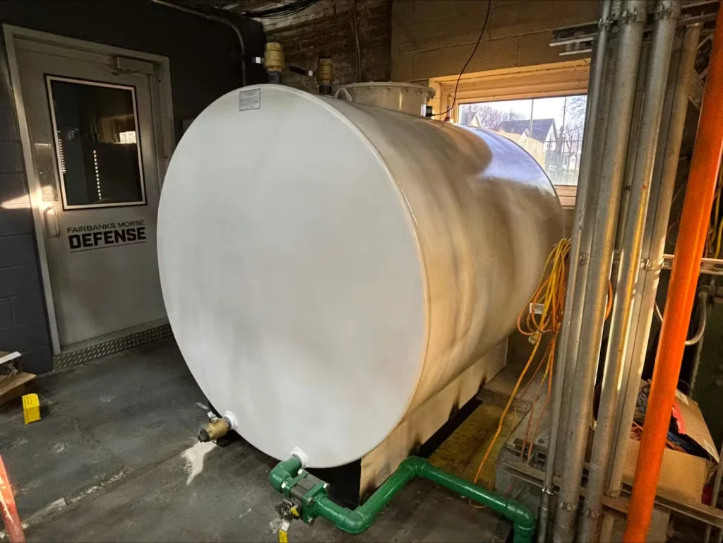 Same utility-room tank freshly painted white, still connected to green piping and a valve at the base; door and window sit behind it.