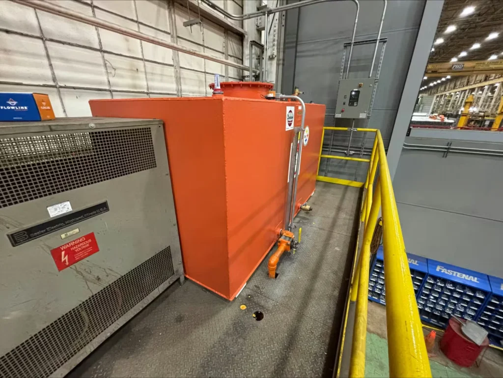 Orange rectangular tank with a top access hatch and orange piping on a metal catwalk next to a yellow railing; an electrical panel and factory interior appear in the background.