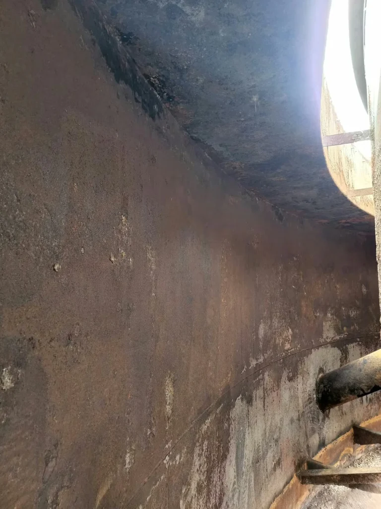 Rusted interior wall of a large cylindrical industrial tank, lit by daylight from an opening at the top right, with a short pipe protruding from the wall.