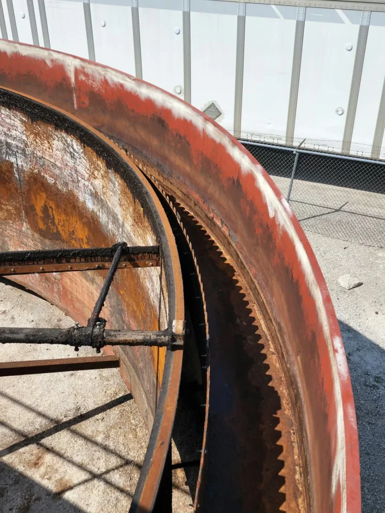 Close-up of a rusted circular steel ring with gear-like teeth and a cross brace, with a chain-link fence and a white trailer in the background.