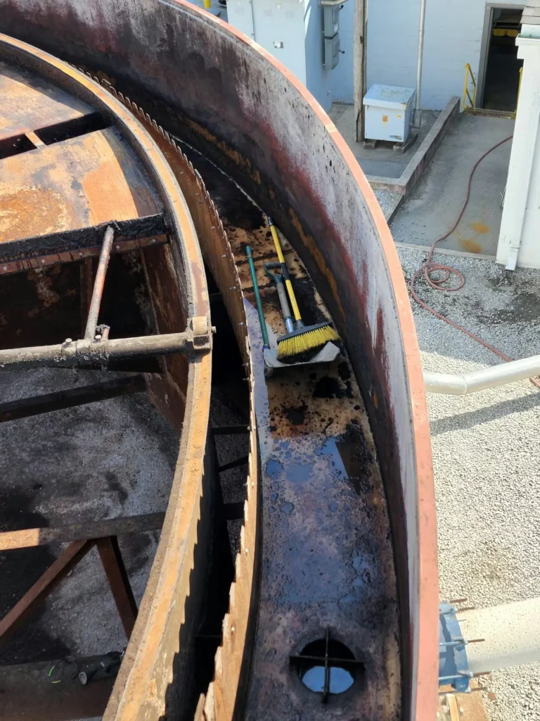 Overhead view into a rusted circular tank section; brooms and a shovel rest in a residue-stained channel along the rim.