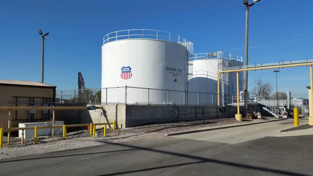 Two large white above-ground fuel storage tanks at an industrial rail facility, one labeled “Diesel Fuel” with a Union Pacific shield logo, beside piping, catwalk stairs, and a gravel yard under a clear blue sky.