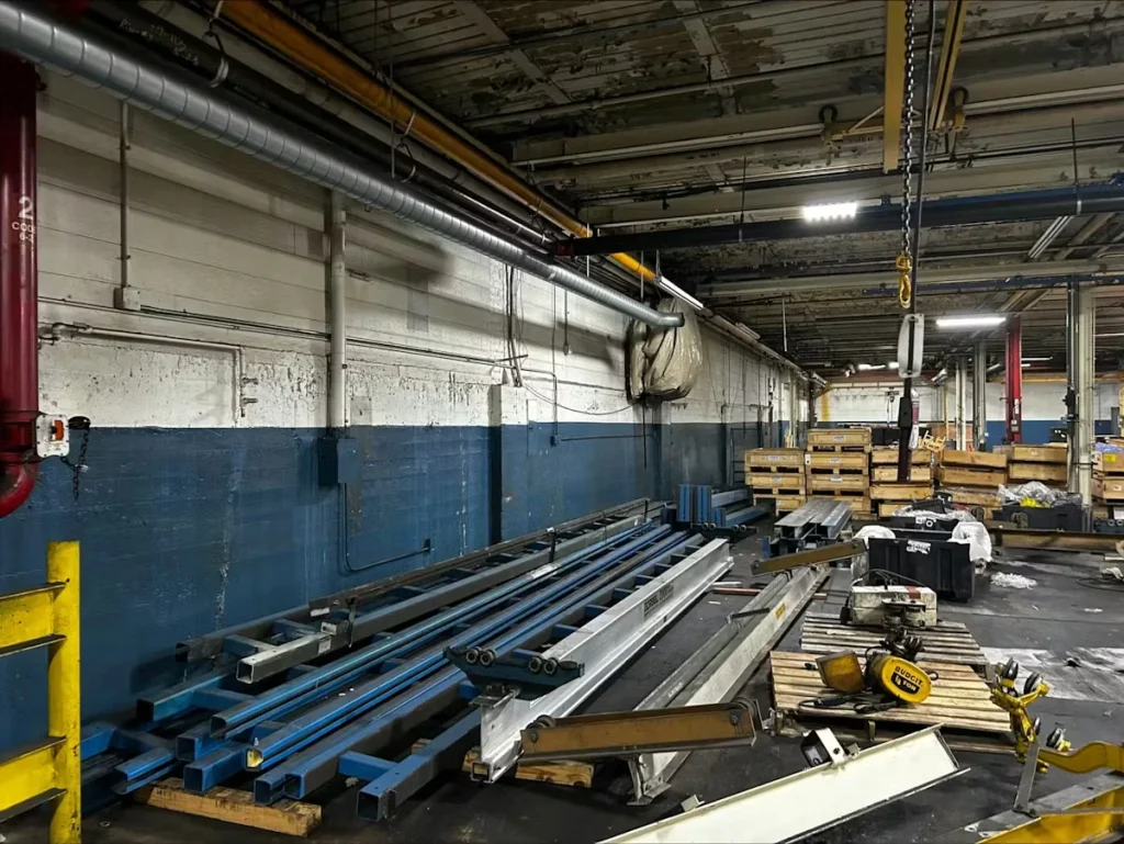 Cluttered industrial warehouse area with a gray wall painted dark blue along the lower half; metal beams and materials lie on pallets, with overhead ductwork, lights, and a hanging hoist chain.