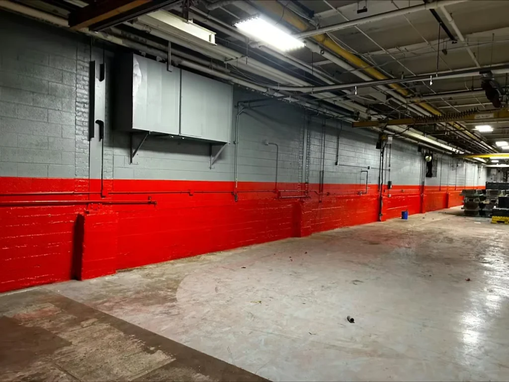 Long, empty industrial bay with a gray cinderblock wall painted bright red along the lower half; exposed pipes and fluorescent lights run across the ceiling above a bare concrete floor.