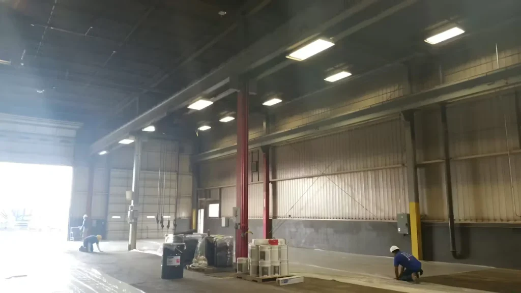 Wide view of a large warehouse interior with high ceilings and bright rectangular overhead lights; plastic sheeting covers parts of the floor while workers kneel near a wall by an open loading-bay door, preparing the area for maintenance or painting.