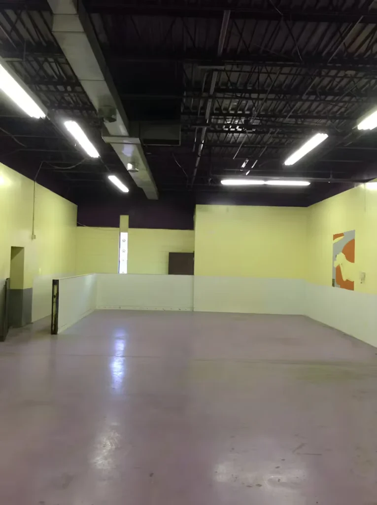 Empty commercial interior with pale yellow walls and a glossy light-purple epoxy floor, lit by long fluorescent fixtures beneath an exposed black ceiling with ducts and beams; a low white half-wall divides the back of the room.