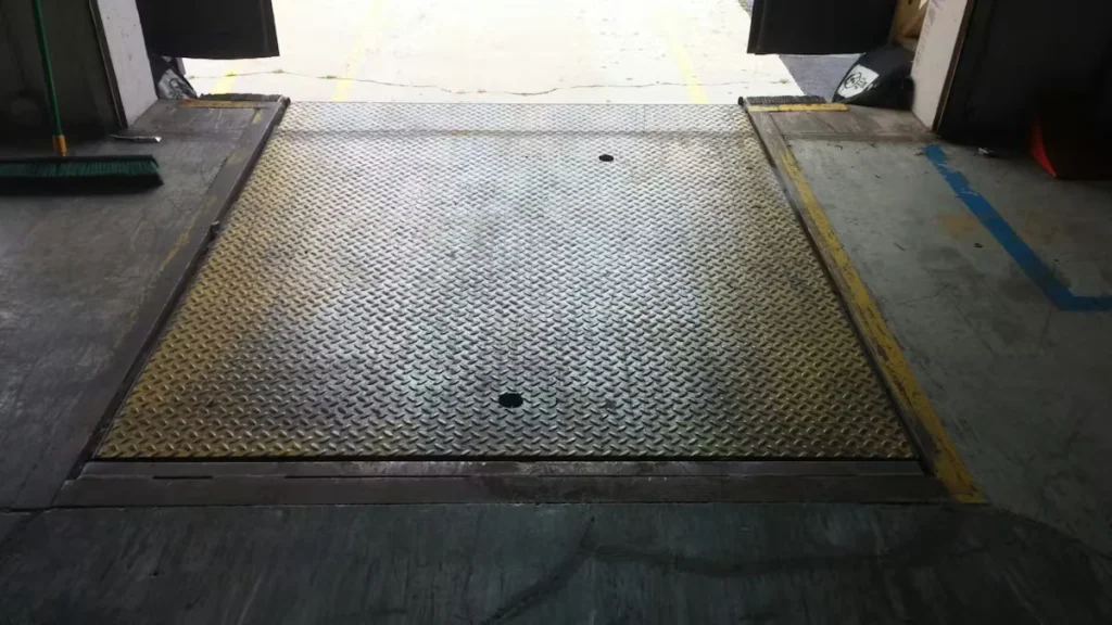 Close-up of a diamond-plate metal dock leveler at a warehouse doorway, bordered by worn concrete and painted safety lines, with daylight visible outside.