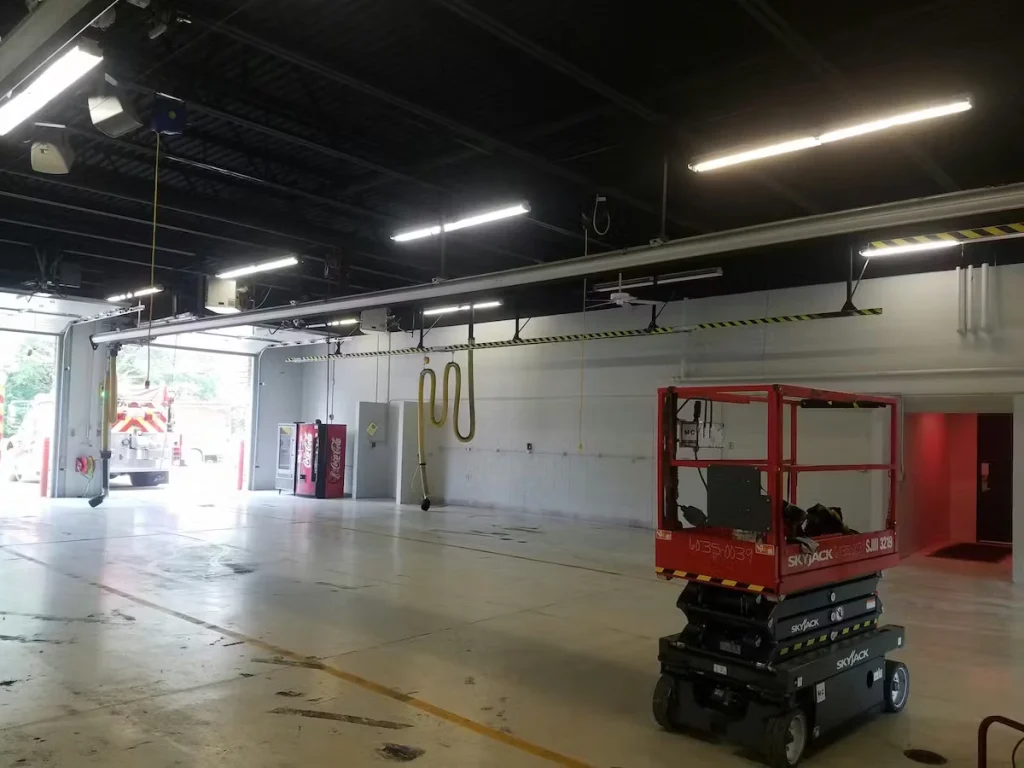 Spacious fire station vehicle bay with a smooth light-colored floor and white walls under a dark ceiling with long fluorescent lights; a red scissor lift is parked in the foreground, large bay doors are open to daylight, and vending machines and hanging hoses line the back wall.