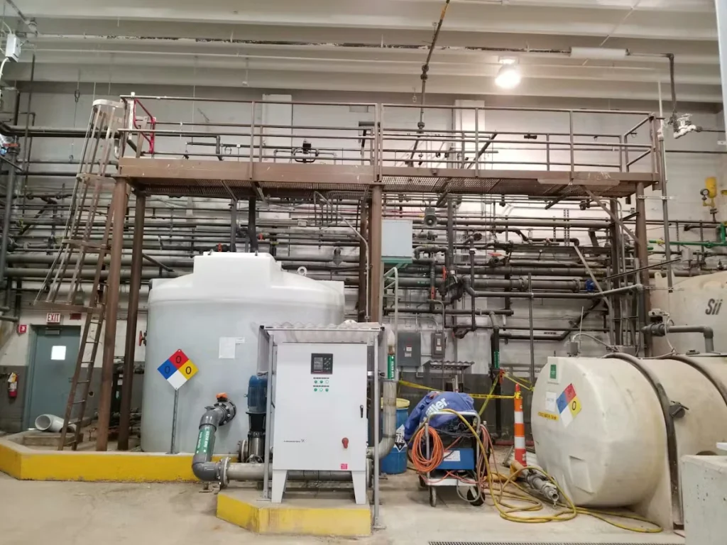 Industrial processing area with large white storage tanks and a control cabinet in front of a wall densely packed with pipes and valves; a metal catwalk with railings spans overhead, with ladders, hoses, and safety markings around the equipment.