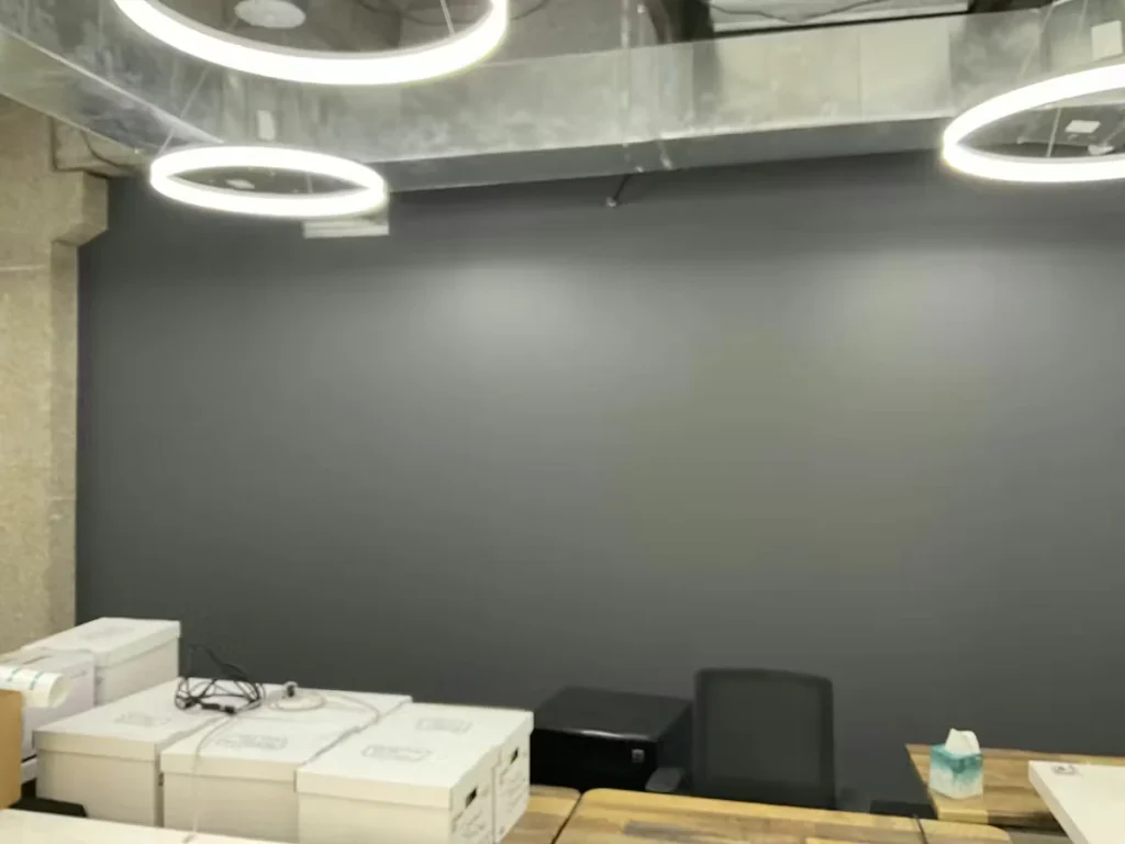 Office reception area with a freshly painted dark gray accent wall, exposed ductwork overhead, and round ring pendant lights; cardboard boxes and a chair sit behind a wood-toned counter in the foreground.