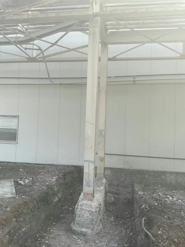 Two vertical steel support columns on a rough concrete footing inside an industrial area, surrounded by excavated dirt trenches; overhead pipe rack framing crosses above, with a light-colored building wall in the background and faint spray-paint markings on the columns.