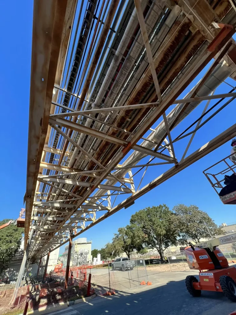 Low-angle view beneath a long elevated pipe rack with multiple large pipes and cable trays, showing rust-streaked steel framing; below are fenced construction areas and an orange boom lift parked on the road at right, with trees and industrial buildings under a clear blue sky.