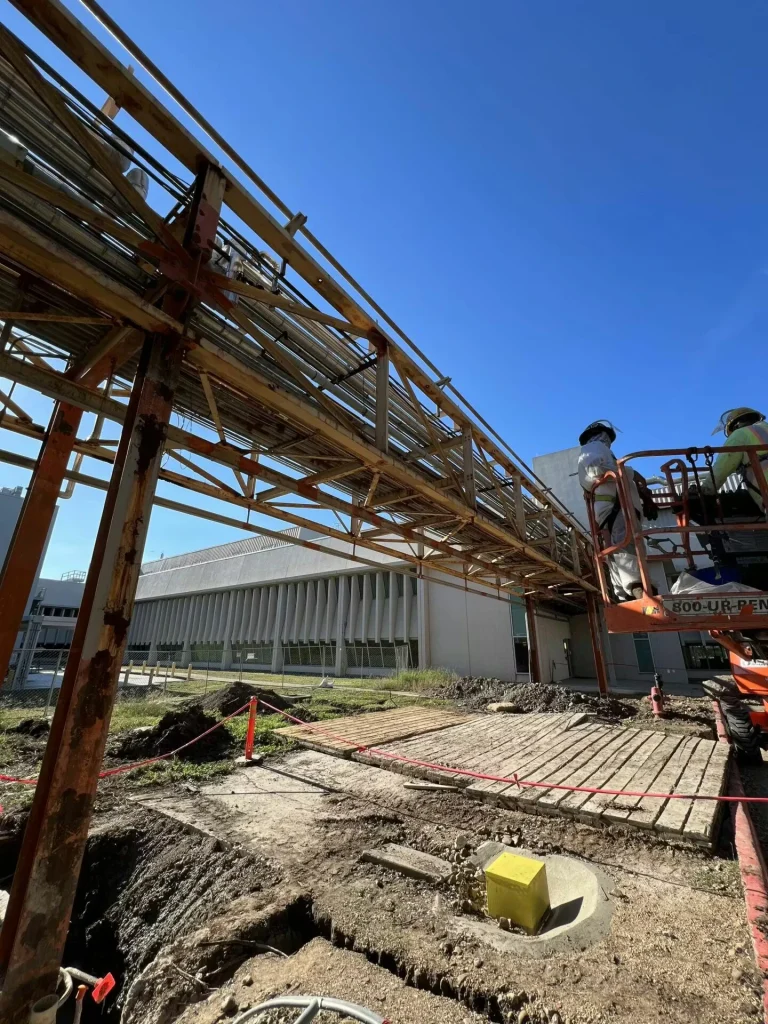Two workers in protective clothing stand on an orange boom lift platform next to a rust-streaked elevated pipe rack; below, an excavated work area with wooden mats and safety rope sits beside a large industrial building under a clear blue sky.