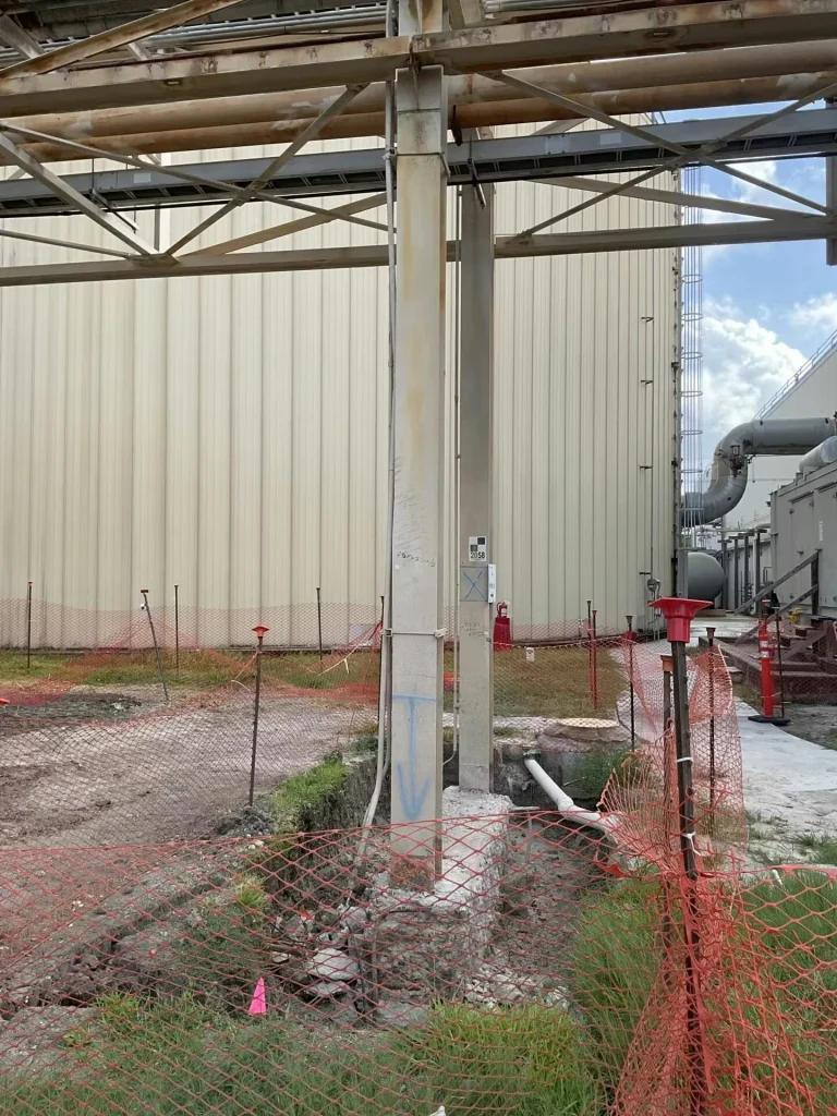Industrial pipe rack columns beside a beige metal-sided building, with orange safety fencing around an open excavation; a narrow trench and exposed white pipe run along the ground near the column bases, and utility equipment and ductwork appear to the right.