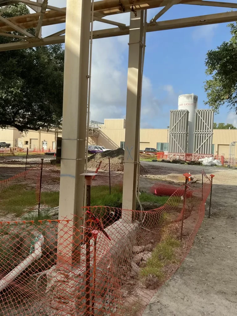 Two tall beige steel support columns beneath a pipe rack at an industrial site, with orange mesh fencing around disturbed ground and temporary piping; in the distance are low industrial buildings and a large vertical tank structure, with trees framing the scene under a partly cloudy blue sky.