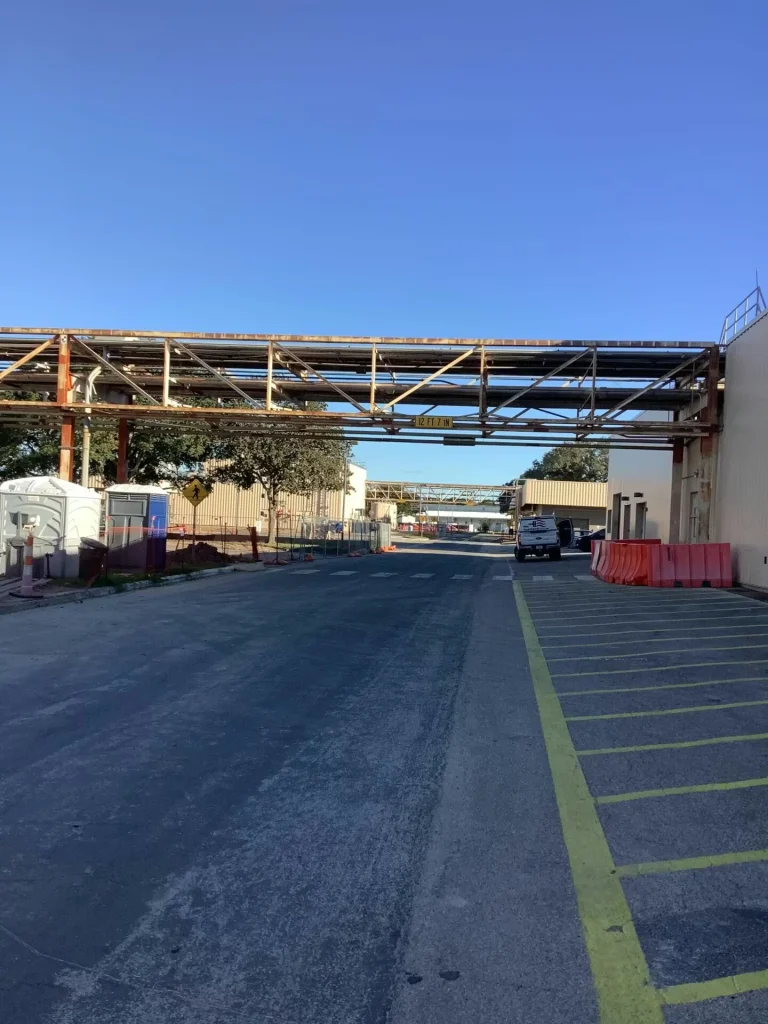 Wide view down an industrial roadway with a metal pipe bridge crossing overhead; portable toilets and construction fencing line the left side, red plastic barriers and marked parking spaces line the right, and a white work van is parked ahead under a clear blue sky.