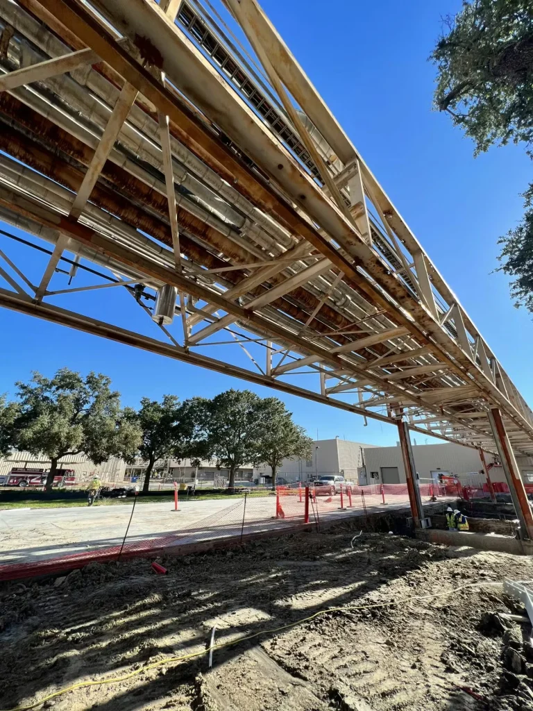 Low-angle view along an elevated pipe rack with multiple lines and cable trays, showing rust-streaked steel members; below, an excavated construction area is bordered by orange safety fencing and cones near an industrial building, with trees and a bright blue sky overhead.