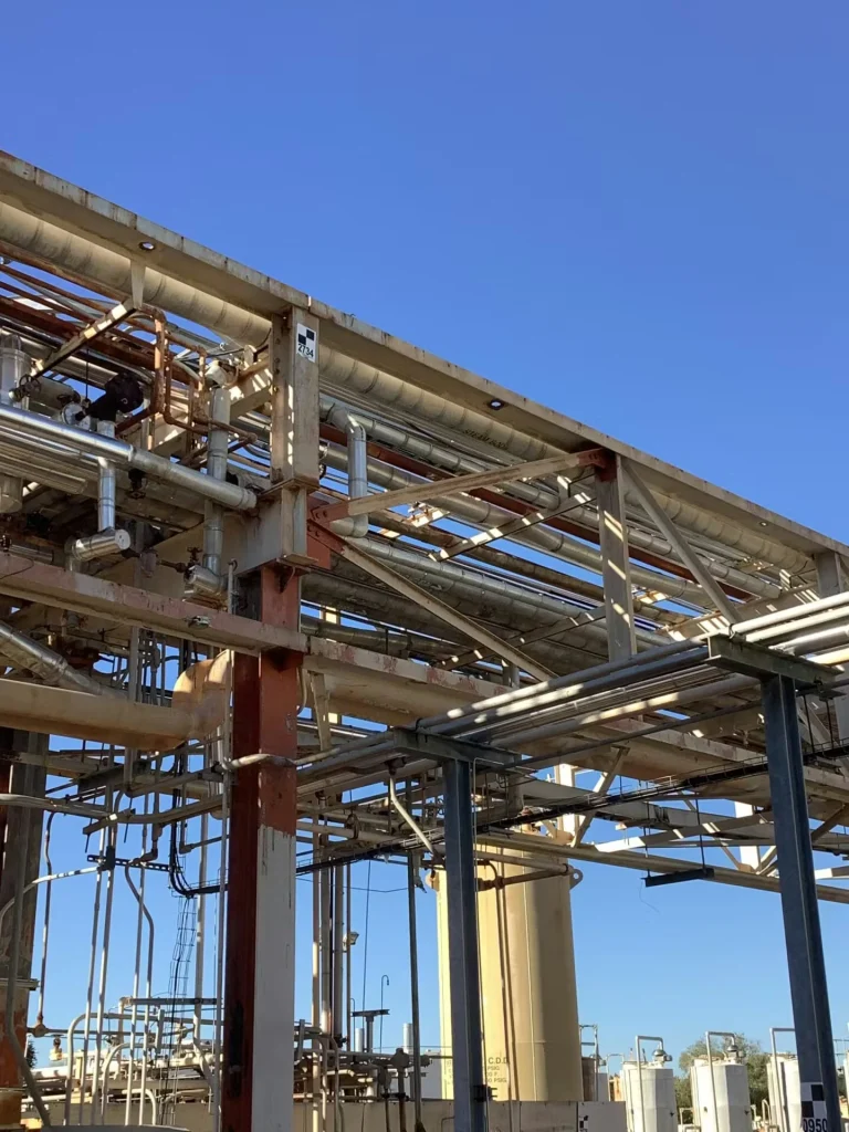 Close-up of an industrial pipe rack structure with intersecting steel beams and numerous pipes running overhead; vertical support columns show rust and patchy paint, and tall cylindrical tanks and process equipment are visible in the background under a clear blue sky.