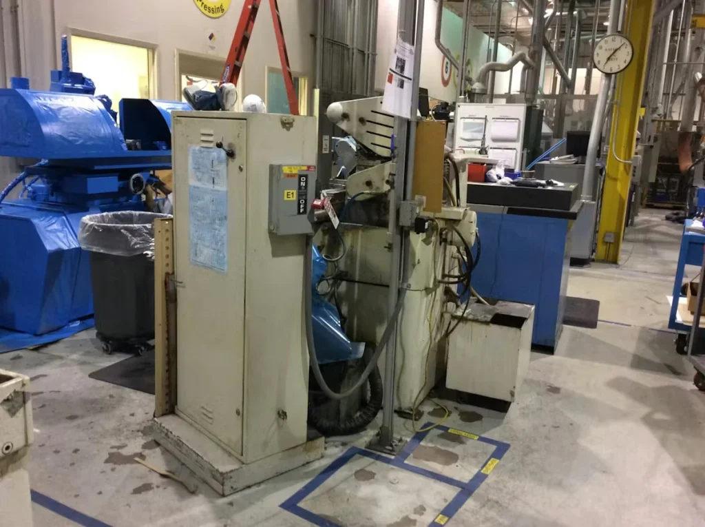 Industrial shop floor with a large piece of machinery and a beige electrical control cabinet in the foreground; blue-painted equipment and workstations sit behind, with taped blue floor markings, overhead piping, and a wall clock visible in the busy factory setting.