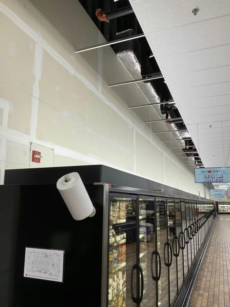 Grocery store freezer aisle under renovation with exposed ceiling grid and unfinished drywall above refrigerated cases