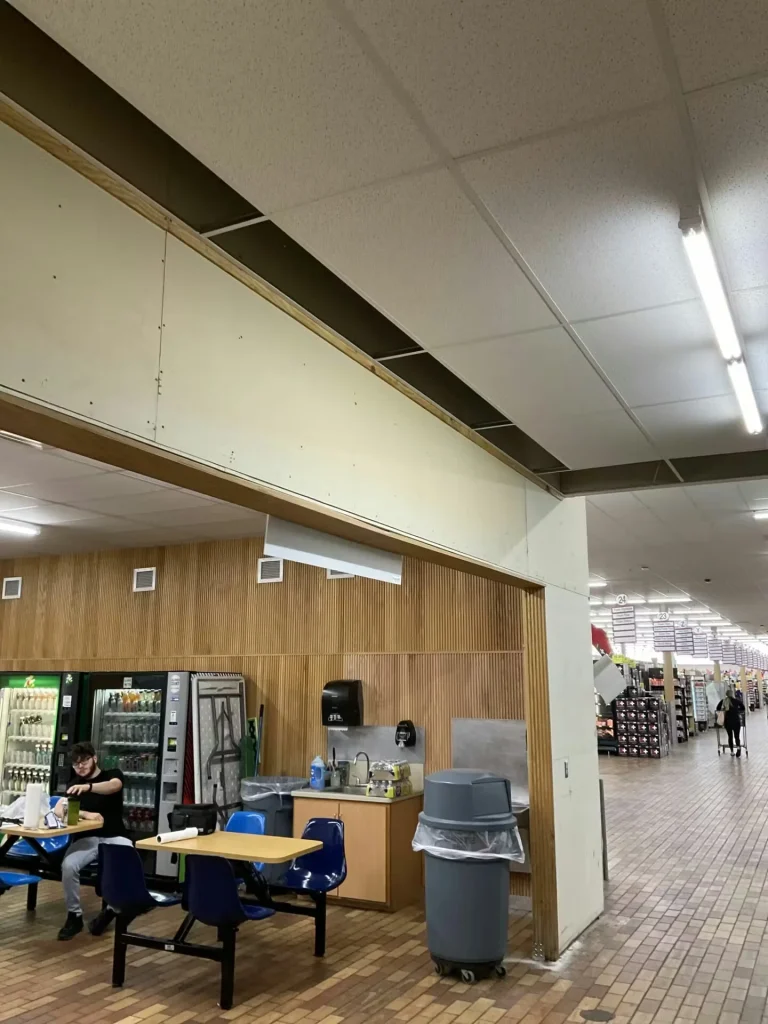 Retail breakroom area during renovation with exposed ceiling sections, seating tables, and open grocery store aisle