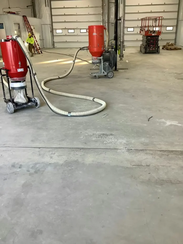 Concrete floor preparation process using industrial vacuum and grinding equipment inside warehouse.