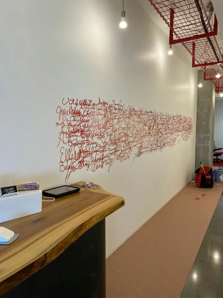 Office hallway wall with red handwritten mural artwork in progress, protective floor covering, and exposed ceiling lighting.