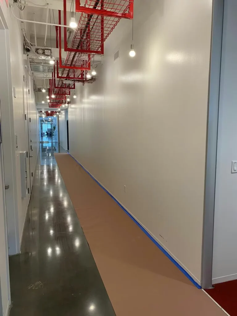Freshly painted office corridor with smooth white walls, red cable trays, polished concrete floor, and floor protection taped in place.