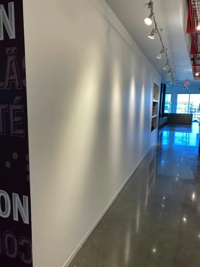 Clean office corridor with newly painted white wall, track lighting, polished concrete floor, and natural light at corridor end.