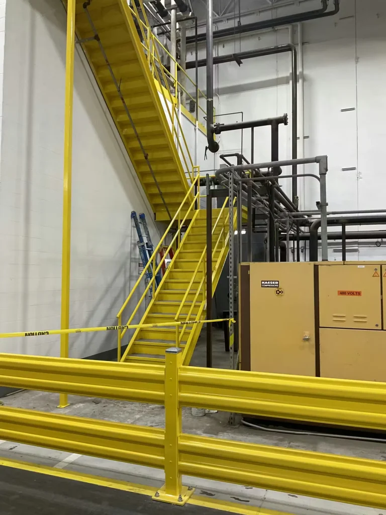 Yellow steel staircase and safety railings inside an industrial plant with exposed piping and equipment