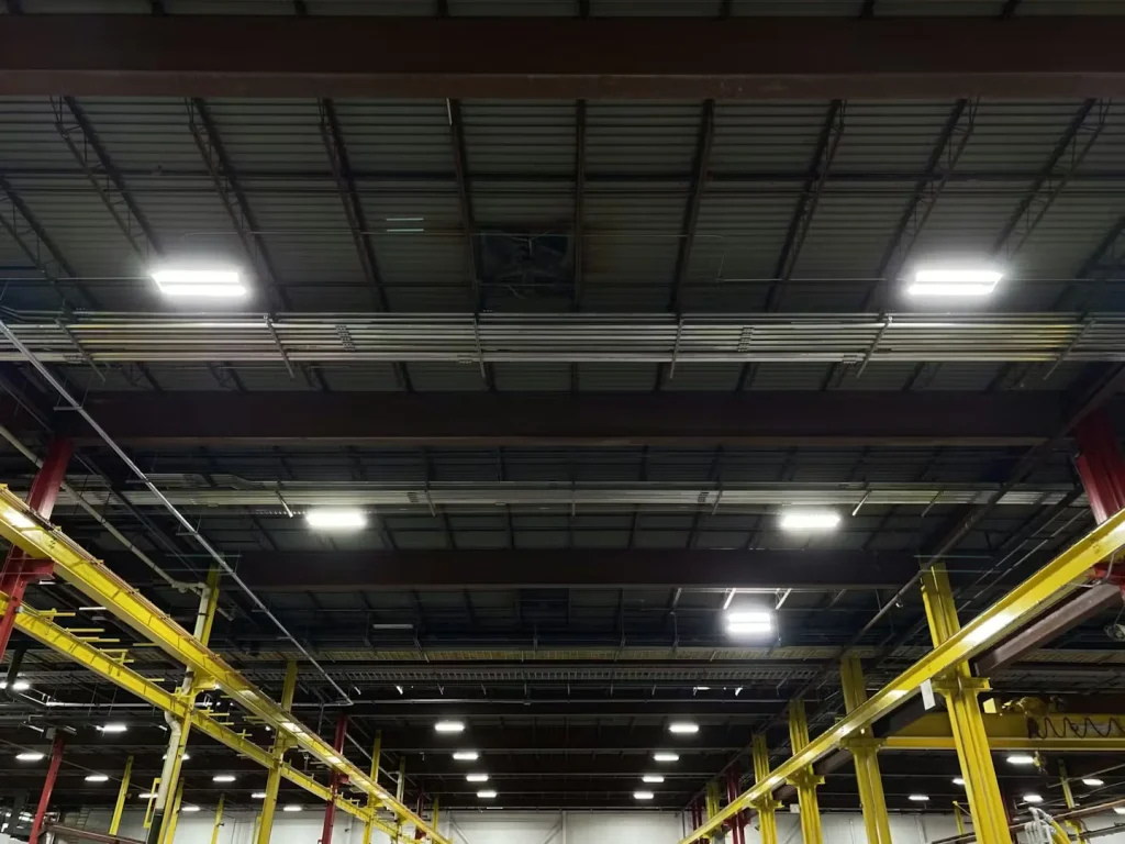 Industrial warehouse ceiling with exposed steel beams, suspended lighting, and conduit lines.
