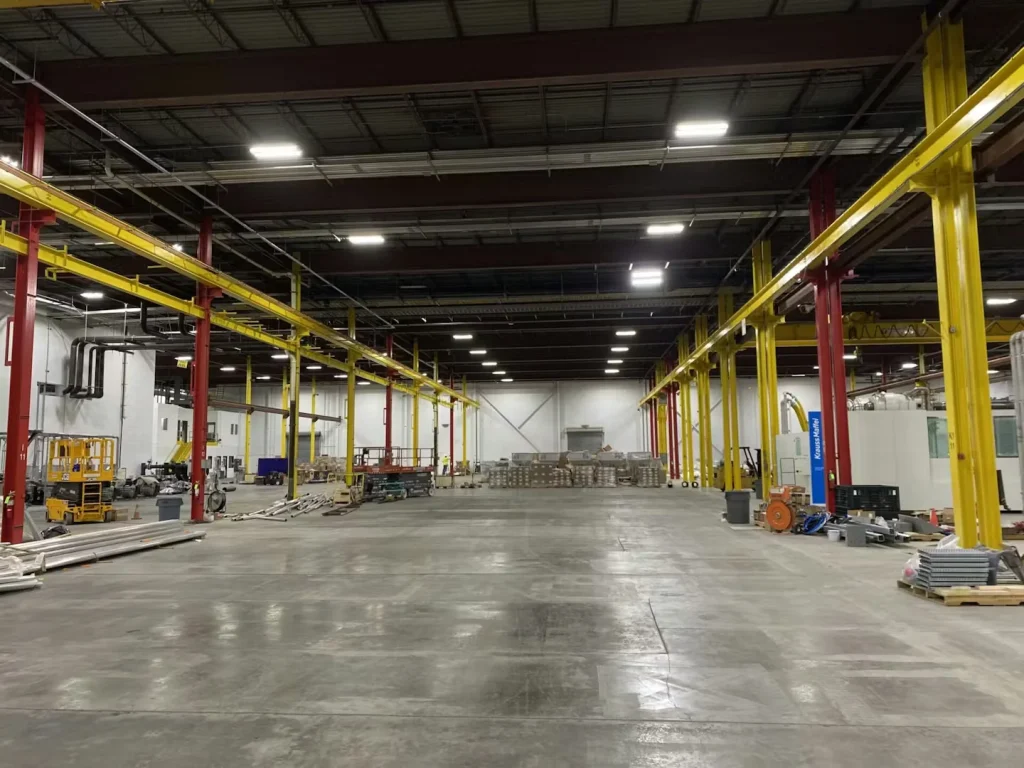 Wide interior of an industrial facility with polished concrete floor, yellow steel columns, overhead crane rails, and construction materials.