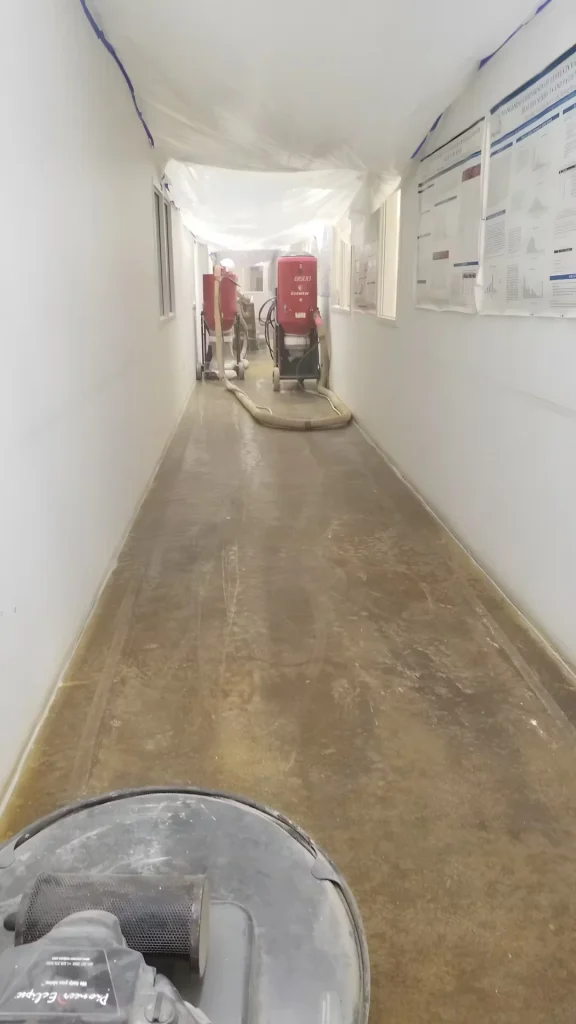 Concrete floor polishing in progress inside a medical laboratory hallway, with dust containment barriers and industrial grinding equipment.