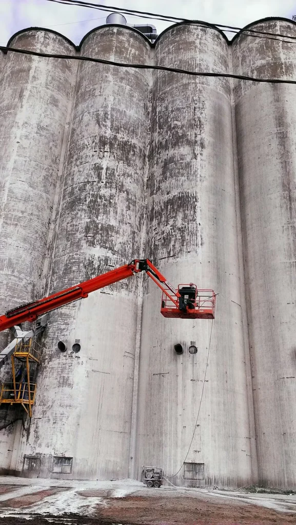 Workers using an aerial lift to prepare and coat large concrete silos at an industrial manufacturing facility