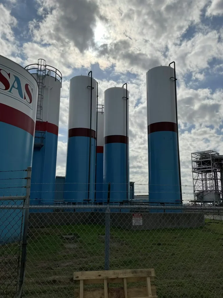 Large industrial storage tanks painted white and blue with red accent bands behind a chain-link fence under a cloudy sky