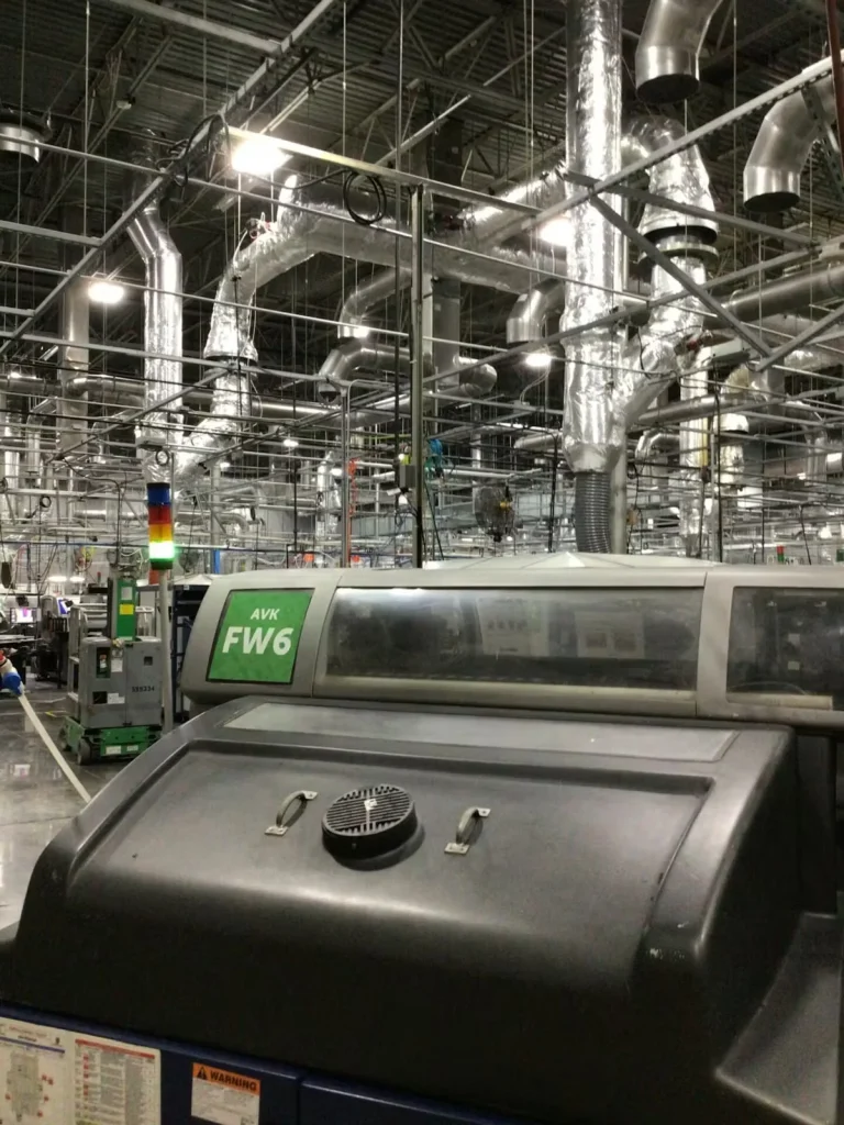 Industrial manufacturing floor with overhead ductwork and ventilation systems, showing a large production machine in the foreground and extensive lint-collection and air-handling infrastructure above, typical of a textile or garment processing facility.
